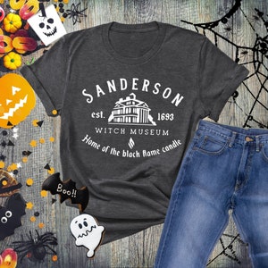 Puede incluir: Una camiseta gris oscuro con un dise&ntilde;o gr&aacute;fico blanco que dice "Sanderson est. 1693 Witch Museum Home of the black flame candle". El gr&aacute;fico tambi&eacute;n incluye una ilustraci&oacute;n en blanco y negro de una casa.