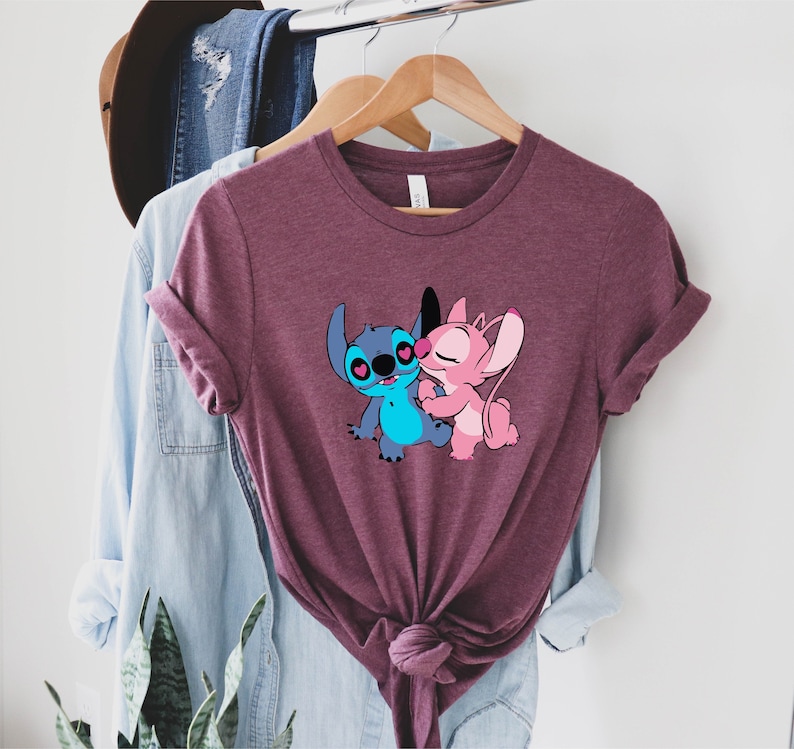 May include: A maroon t-shirt with a graphic of Stitch and Angel from Lilo & Stitch. Stitch is blue and Angel is pink. They are hugging and have hearts for eyes.
