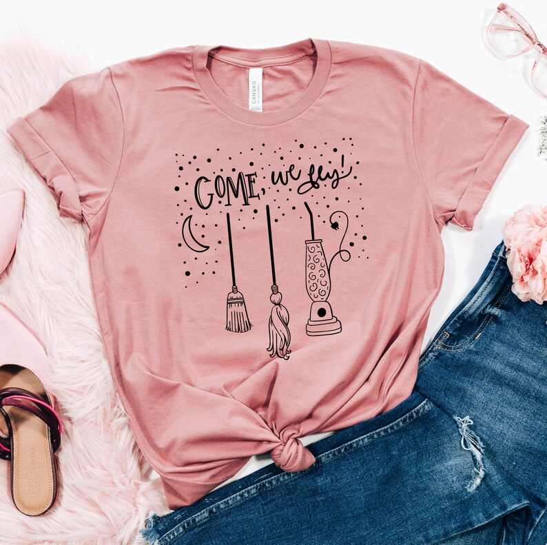 May include: A pink t-shirt with the text "COME, we fly!" in black lettering. The design includes a crescent moon, three broomsticks, and a black silhouette of a lava lamp.