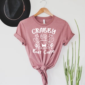 May include: A pink t-shirt with a white graphic of a crab with the text "Crabby But Cute".