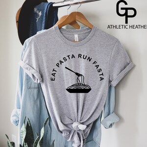 Może przedstawiać: Szary T-shirt z napisem "Eat Pasta Run Fasta" w czarnym kolorze. Na środku koszulki znajduje się czarno-biała ilustracja widelca obracającego spaghetti.