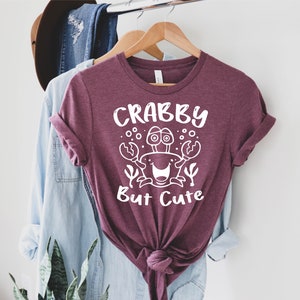 May include: A maroon t-shirt with a white graphic of a crab with the text "Crabby But Cute".