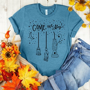 May include: A teal blue t-shirt with the text "COME, we fly!" in black lettering. The design includes a crescent moon, three broomsticks, and a lava lamp.