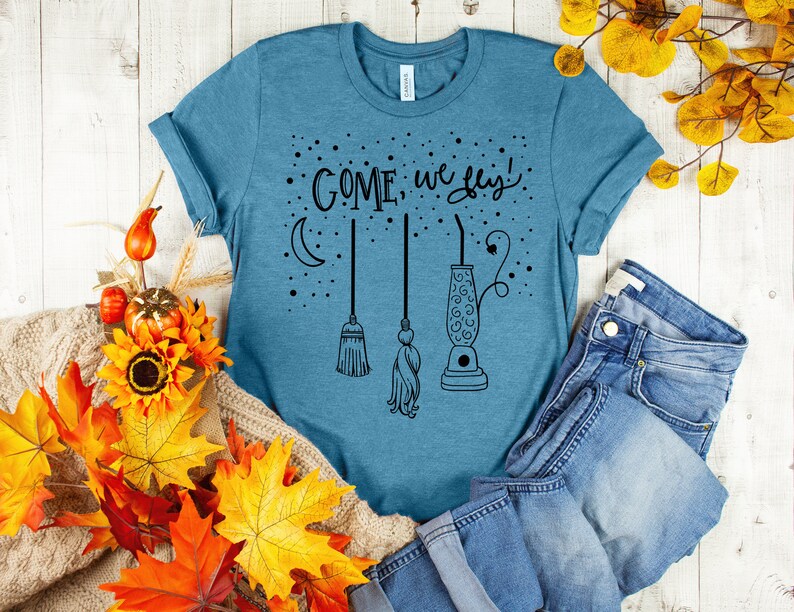 May include: A teal blue t-shirt with the text "COME, we fly!" in black lettering. The design includes a crescent moon, three broomsticks, and a lava lamp.