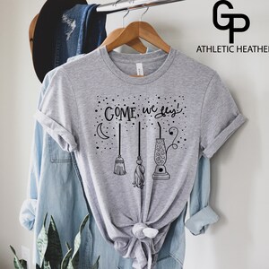 May include: Gray t-shirt with a black graphic of broomsticks and a cauldron with the text "COME, we fly!" printed on it.