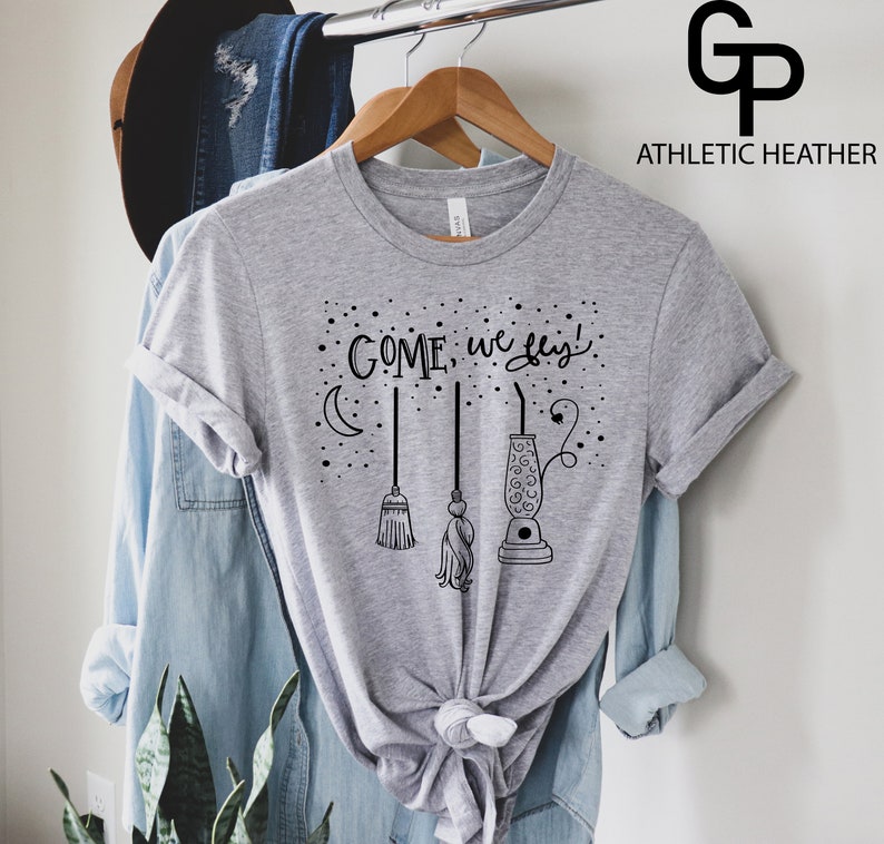 May include: Gray t-shirt with a black graphic of broomsticks and a cauldron with the text "COME, we fly!" printed on it.