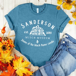Puede incluir: Una camiseta azul turquesa con un dise&ntilde;o gr&aacute;fico blanco que dice "Sanderson est. 1693 Witch Museum Home of the black flame candle". El gr&aacute;fico tambi&eacute;n incluye una silueta blanca de una casa.
