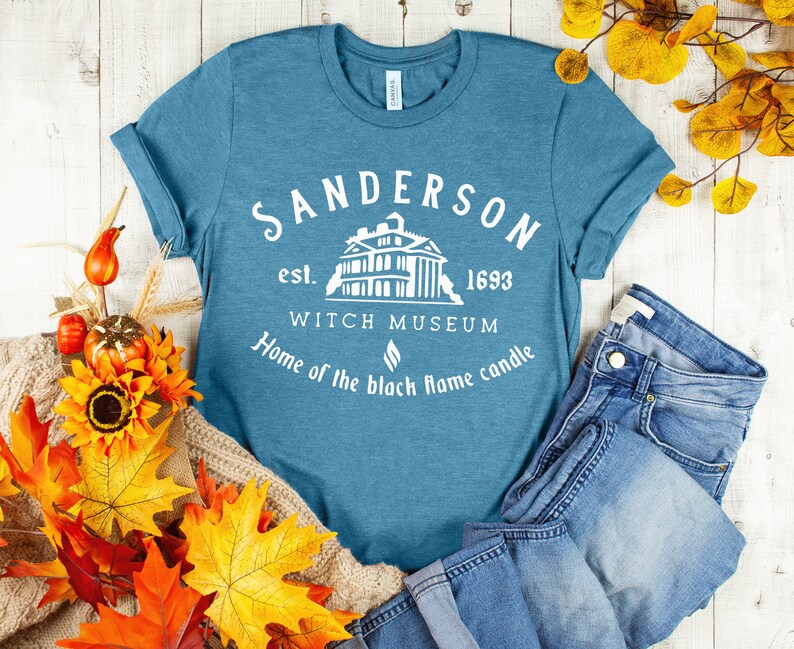 Puede incluir: Una camiseta azul turquesa con un dise&ntilde;o gr&aacute;fico blanco que dice "Sanderson est. 1693 Witch Museum Home of the black flame candle". El gr&aacute;fico tambi&eacute;n incluye una silueta blanca de una casa.