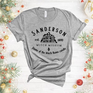Puede incluir: Una camiseta gris con un dise&ntilde;o gr&aacute;fico negro que presenta el texto "Sanderson est. 1693 Witch Museum Home of the black flame candle".