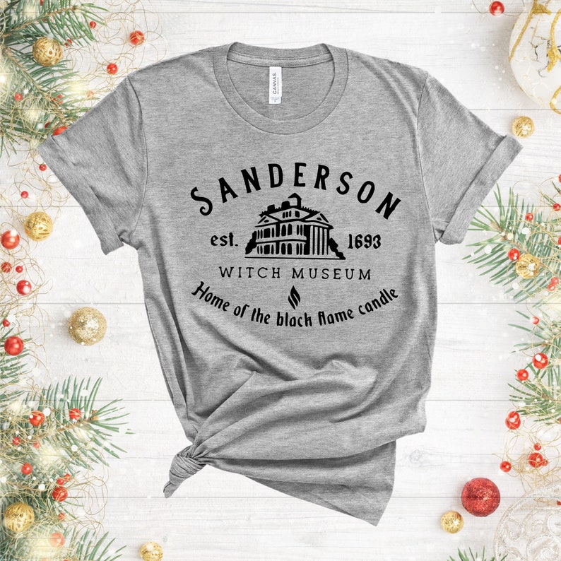 Puede incluir: Una camiseta gris con un dise&ntilde;o gr&aacute;fico negro que presenta el texto "Sanderson est. 1693 Witch Museum Home of the black flame candle".