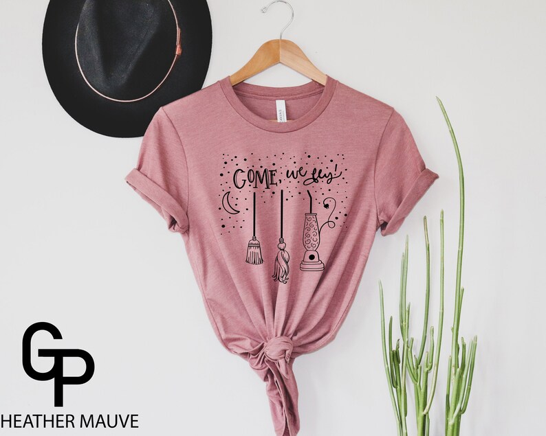 May include: A heather mauve t-shirt with the text "COME, we fly!" in black lettering. The text is accompanied by a crescent moon, a broom, and a lantern.