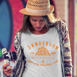 Puede incluir: Camiseta blanca con un dise&ntilde;o gr&aacute;fico naranja que representa una casa con el texto "Sanderson est. 1693 Witch Museum Home of the black flame candle".