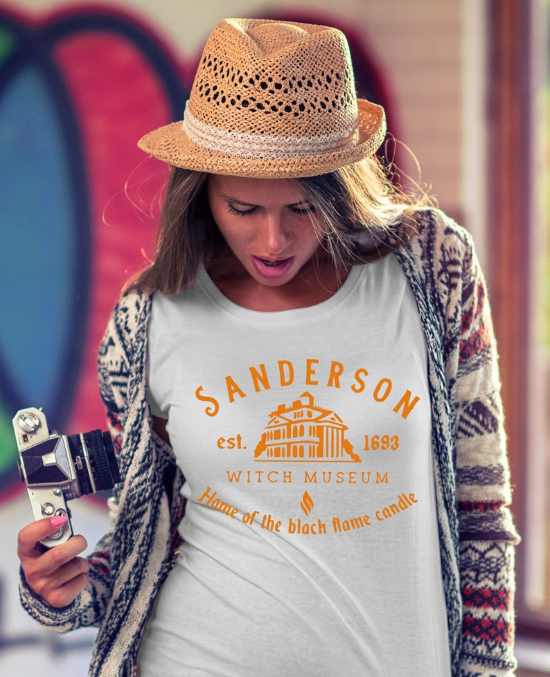 Puede incluir: Camiseta blanca con un dise&ntilde;o gr&aacute;fico naranja que representa una casa con el texto "Sanderson est. 1693 Witch Museum Home of the black flame candle".