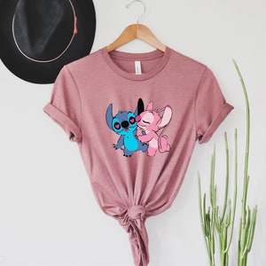 May include: A pink t-shirt with a graphic of Stitch and Angel from Lilo & Stitch. The characters are hugging and have hearts for eyes.