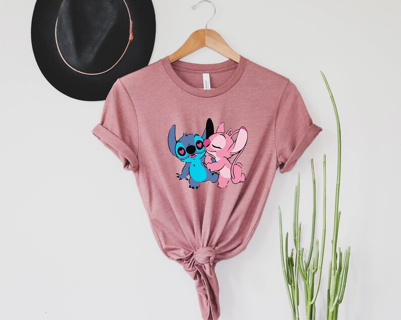 May include: A pink t-shirt with a graphic of Stitch and Angel from Lilo & Stitch. The characters are hugging and have hearts for eyes.