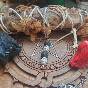 May include: A silver chain necklace with skull and lava bead pendants, a red stone, and a black stone are displayed on a wooden surface with a pentagram design. A bundle of dried sage is also present.