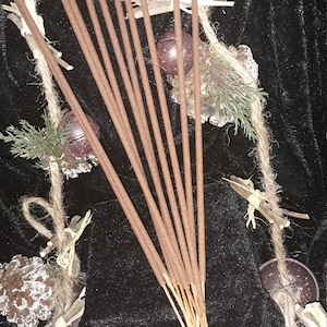 May include: A bundle of brown incense sticks with a natural wood scent. The sticks are arranged in a fan shape and are tied together with twine. The incense sticks are surrounded by decorative elements, including pine cones, bells, and twigs.