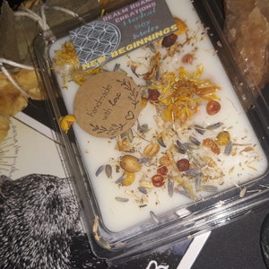 Könnte beinhalten: Ein weißer Wachsschmelz mit getrockneten Blumen und Kräutern in einem durchsichtigen Plastikbehälter. Das Etikett lautet "Realm Roar Creations Herbal Soy Melts New Beginnings" und "Handmade with Love".