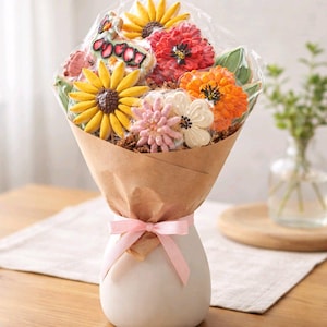 May include: A floral-themed bouquet of decorated sugar cookies, wrapped in brown paper and tied with a pink ribbon. The cookies are shaped like sunflowers, daisies, and other flowers. The bouquet is displayed in a white vase.