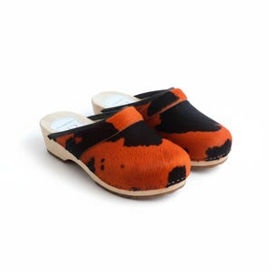 May include: A pair of clogs with a wooden sole and a black and orange patterned upper. The clogs have a black strap across the top of the foot. The wooden sole is approximately 1 inch thick.