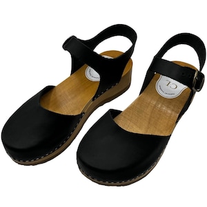 Puede incluir: Zuecos de cuero negro con suela de plataforma de madera y una correa de tobillo con hebilla. Los zuecos tienen una puntera redondeada y un detalle de costura blanca alrededor de la suela.