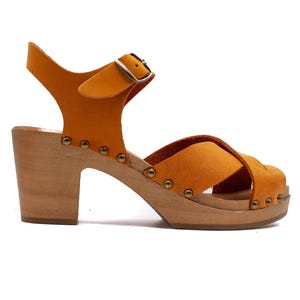 Puede incluir: Sandalias de tacón de cuero naranja con cierre de hebilla y plataforma de madera. Las sandalias tienen un diseño de tiras cruzadas y están adornadas con pequeños tachuelas metálicas.