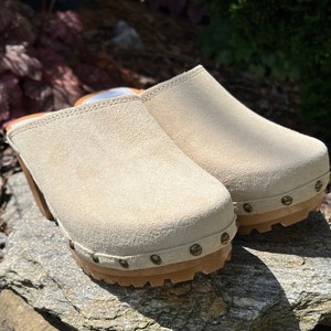 Peut inclure: Une paire de sabots en daim beige avec une semelle en bois et une semelle en caoutchouc. Les sabots ont un bout arrondi et sont ornés de petits clous métalliques autour de la semelle. Les chaussures sont posées sur une surface en pierre naturelle.