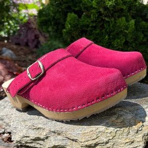 Puede incluir: Un par de zuecos de ante burdeos con una hebilla. Los zuecos tienen una base de madera y están sobre una pila de piedras.