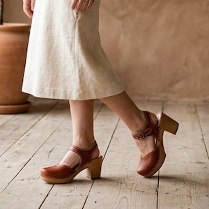 Puede incluir: Un vestido de color crema, de manga corta y largo hasta la rodilla, con escote redondo y cuerpo entallado que se ensancha en la falda, combinado con zuecos de cuero marrón con suela de madera y una tira con hebilla sobre el empeine.