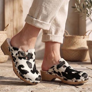 May include: A pair of slip-on clogs featuring a black and white cow print pattern on the upper, a brown wooden sole, and a metallic buckle strap across the top. The clogs are constructed with visible metal studs securing the leather to the wooden base.