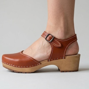 Puede incluir: Un zueco de cuero marrón con punta redondeada, suela de madera clara y una correa de tobillo ajustable con hebilla de metal. El zapato presenta costuras blancas visibles a lo largo de los bordes y una suela de goma marrón con textura.