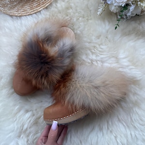 May include: A pair of brown leather clogs with a thick wooden sole, adorned with fluffy, multi-toned fur trim. The fur features shades of brown, tan, and grey. The clogs are set on a white, fluffy surface.