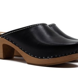 Puede incluir: Zuecos de cuero negro con tacón de madera y un detalle de costura blanca alrededor del borde.