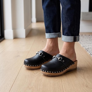 A person stands on a light wood floor, wearing dark blue cuffed jeans and black leather clogs with silver-toned metal buckles and wooden soles.