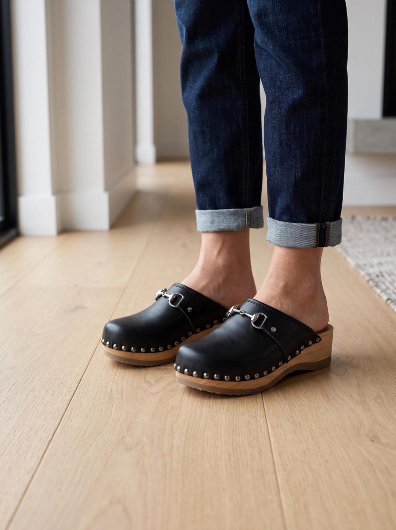 A person stands on a light wood floor, wearing dark blue cuffed jeans and black leather clogs with silver-toned metal buckles and wooden soles.