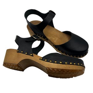 May include: Black leather clogs with a wooden heel and gold studs. The clogs have an ankle strap with a buckle.