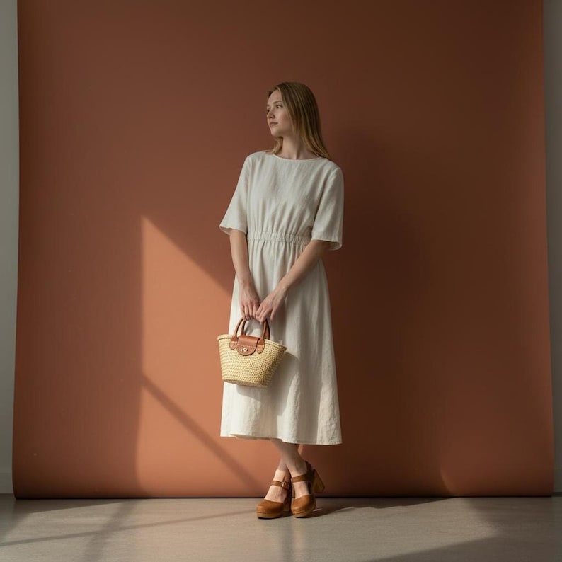 Woman wearing handmade brown leather wooden clogs with ankle strap and block wood heel, styled with cream linen dress against warm neutral background.