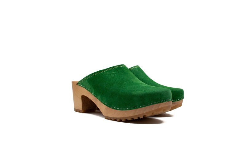 Puede incluir: Zuecos de ante verde con tac&oacute;n y plataforma de madera. Los zuecos tienen un detalle de costura blanca alrededor del borde.