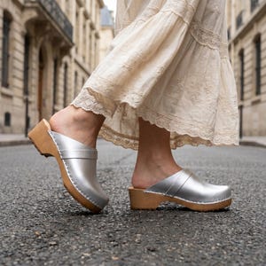 Puede incluir: Un par de zuecos sin cordones con una parte superior de color plateado metálico y una suela de madera natural clara. Los zuecos tienen la punta redondeada, una tira ancha en el empeine y un tacón bajo, combinados con una falda color crema con encaje.