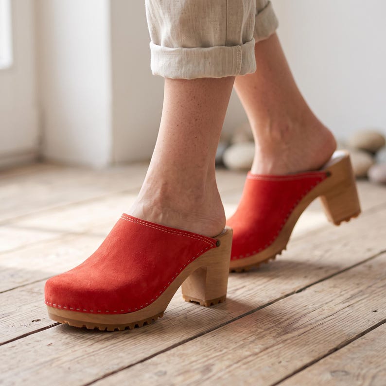 A person with light skin and light-colored pants walks on a light wooden floor, wearing red suede clogs with light wooden soles and heels.