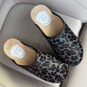 May include: A pair of clogs with a wooden sole and a leopard print upper. The clogs have a black trim and a label that reads "CLOGSLAND.COM". The clogs are made of natural leather.