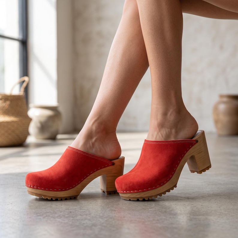 A person with light skin and bare legs stands on a light-colored floor, wearing red suede clogs with light wooden soles and heels.