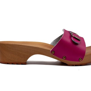 May include: A pair of pink leather clogs with a wooden platform and a buckle detail. The clogs have a low heel and a textured sole.