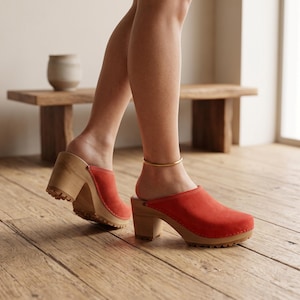 A person with light skin and bare legs, wearing a gold ankle bracelet, walks on a light wooden floor, wearing red suede clogs with light wooden soles and heels.