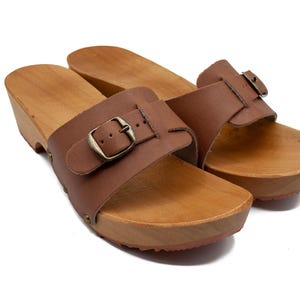 A pair of brown leather clogs with wooden soles and buckled straps, viewed from above and slightly to the front.