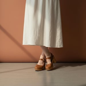 Woman wearing handmade brown leather wooden clogs with ankle strap and block wood heel, styled with cream linen dress against warm neutral background.