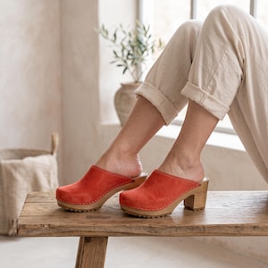 A person with light skin and light-colored pants sits on a wooden bench, wearing red suede clogs with light wooden soles and heels.