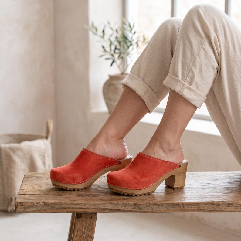 A person with light skin and light-colored pants sits on a wooden bench, wearing red suede clogs with light wooden soles and heels.