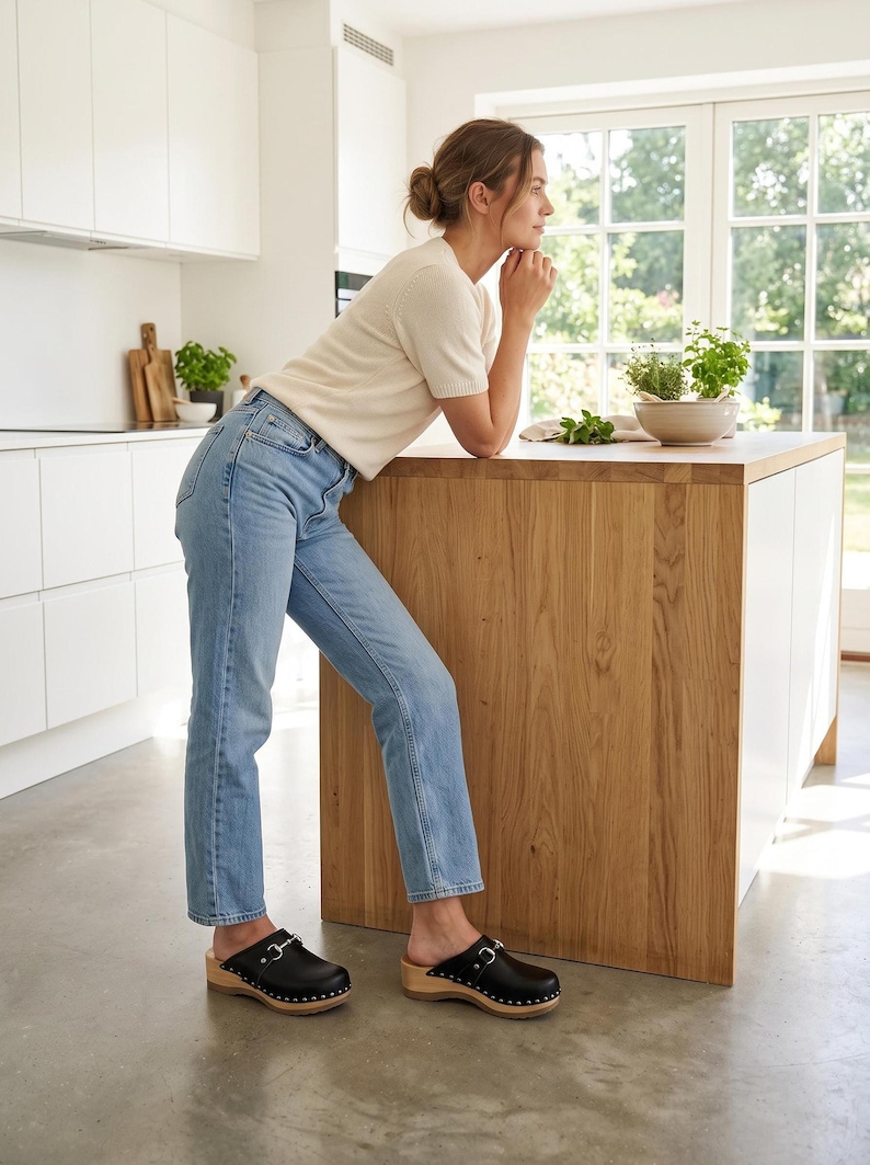 A woman in a white sweater and blue jeans stands in a kitchen, wearing black leather clogs with silver-toned metal buckles and wooden soles, leaning on a light wood island.