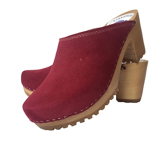 May include: Red suede clogs with a wooden heel and a white stitched trim. The clogs have a textured sole.